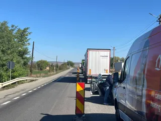 Un camion alb și un van gri stau pe marginea unui drum asfaltat lângă un con de circulație galben și roșu sub un cer senin.