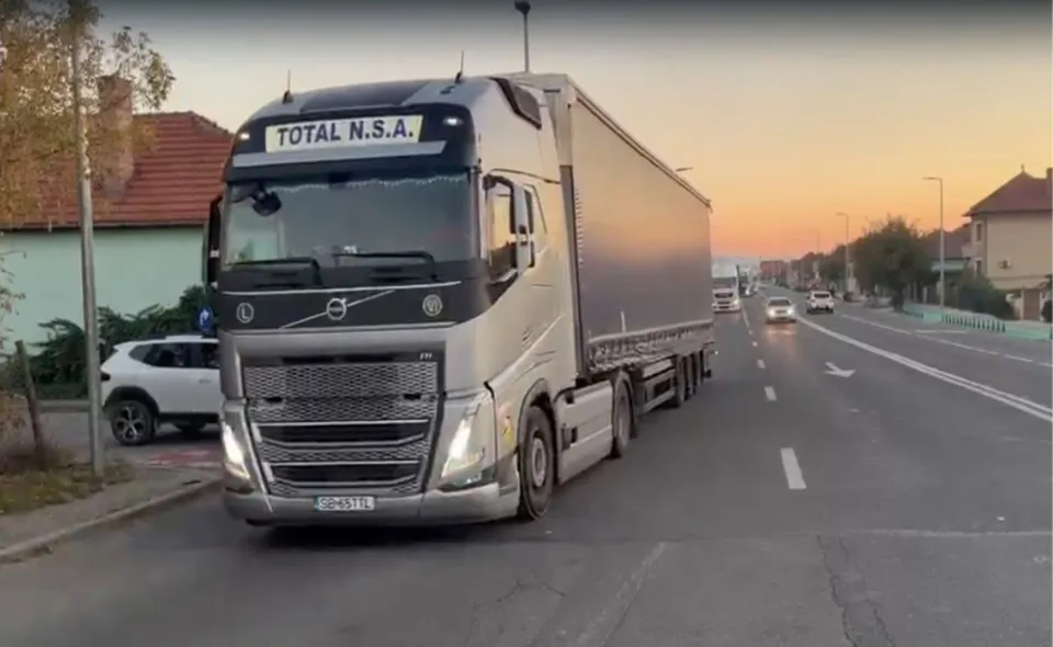 Un camion mare de culoare gri cu inscripția TOTAL N.S.A. circulă pe un drum asfaltat lângă o clădire și un autoturism alb.