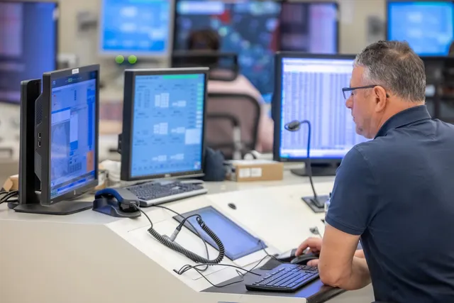 Un bărbat cu ochelari stă la un birou cu mai multe monitoare, un tastatură și un mouse într-un birou iluminat.