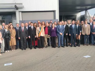 Un grup de persoane îmbrăcate în haine formale stă în rând în fața unei clădiri industriale cu uși metalice gri.