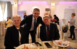 Un bărbat într-un costum negru stă între o femeie cu sacou negru și un bărbat cu ochelari și costum negru la o masă cu farfurii.
