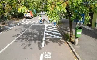 Trei persoane în veste reflectorizante lucrează pe un drum asfaltat lângă marcaje albe, în timp ce un camion portocaliu este parcat în fundal.
