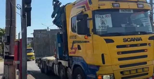 Un camion galben de mare tonaj este parcat pe un drum, având un semn alb cu textul STOP afișat pe parbrizul frontal.