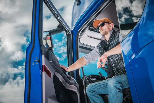 Un bărbat cu basetă și vestă cu carouri stă în cabina unui camion albastru, uitându-se în lateral.