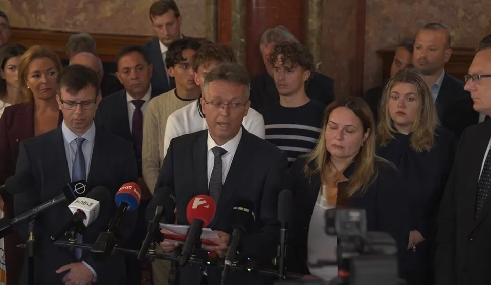 Un bărbat cu ochelari și cravată deschide un document în fața mai multor microfoane, fiind înconjurat de un grup de persoane într-o încăpere interioară.