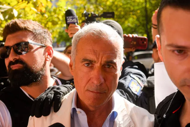 Un bărbat cu părul scurt și alb gri este încadrat de alți bărbați, unul purtând ochelari de soare și altul având o mână pe umărul său.