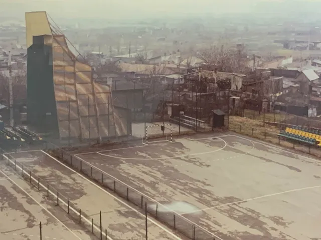 O structură verticală înaltă cu suprafețe colorate este situată lângă un teren de sport delimitat de un gard în fața unor clădiri urbane.