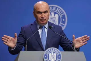 Un bărbat cu costum și cravată stă în spatele unui podium cu microfoane, având mâinile ridicate în lateral pe un fundal albastru.