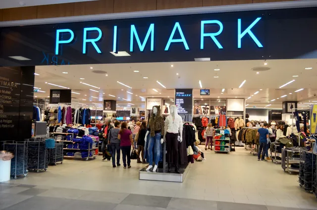 Intrarea unui magazin de haine cu un semn luminos albastru deasupra, prezentând manechine și rafturi cu îmbrăcăminte în interiorul unei încăperi luminoase.