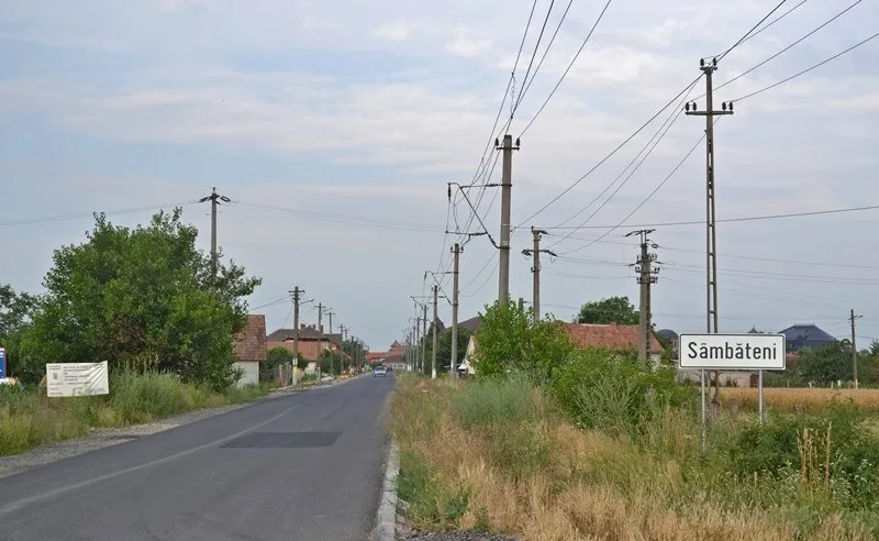 O stradă asfaltată cu linii de utilități și un panou indicativ alb pe care scrie Sâmbăteni se află lângă vegetație și case.