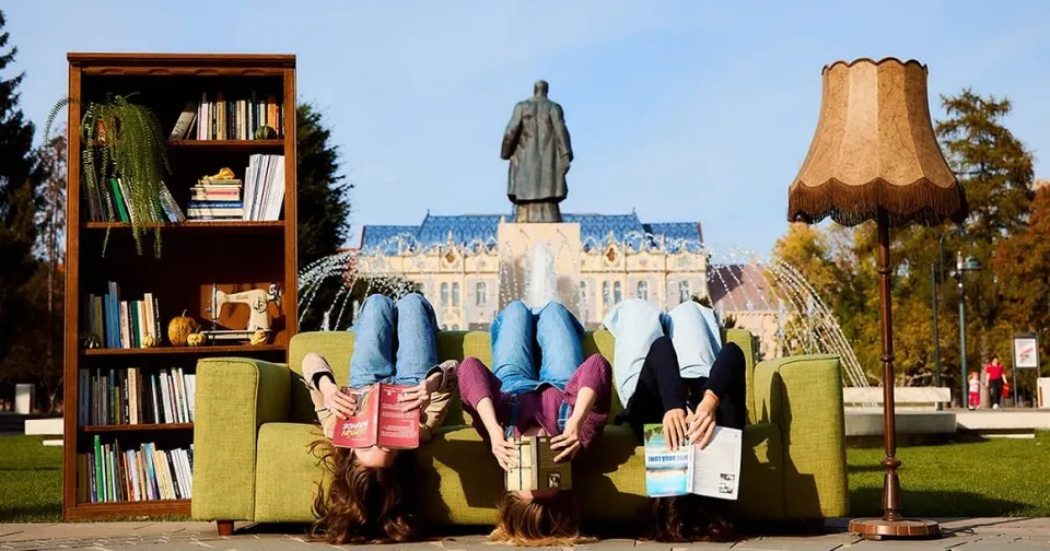 Trei persoane stau întoarse pe spate pe o canapea verde, citind cărți, lângă o bibliotecă și o lampă de podea.