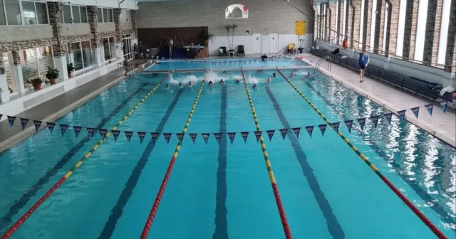 O piscină interioară cu linii de culoare albastră și marcaje de culoare galbenă și roșie, cu câteva persoane în apă la capătul bazinului.
