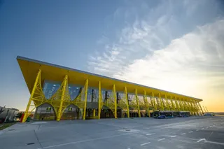 O clădire modernă cu fațadă de sticlă și structură metalică galbenă se află pe o suprafață asfaltată sub un cer cu nori la apus.
