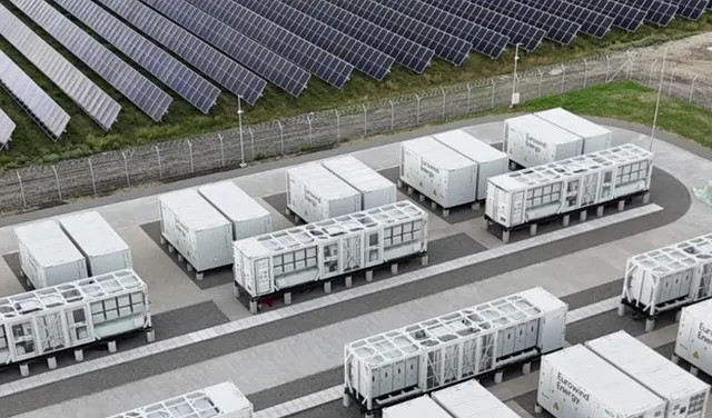 Rânduri de panouri solare sunt instalate pe un teren verde, lângă containere albe de stocare a energiei dispuse pe platforme de beton.