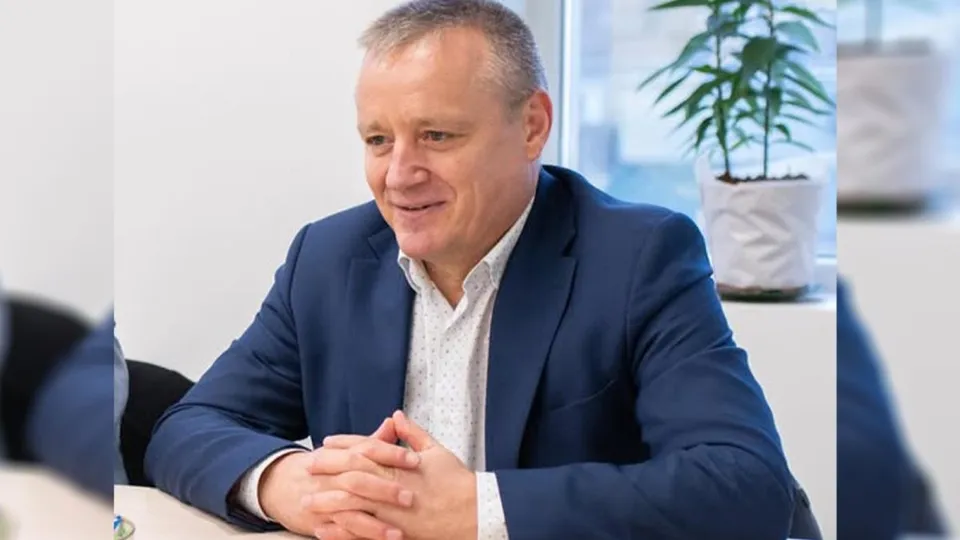 Un bărbat cu părul scurt, purtând un sacou albastru și o cămașă albă cu puncte, stă la o masă cu mâinile încrucișate.