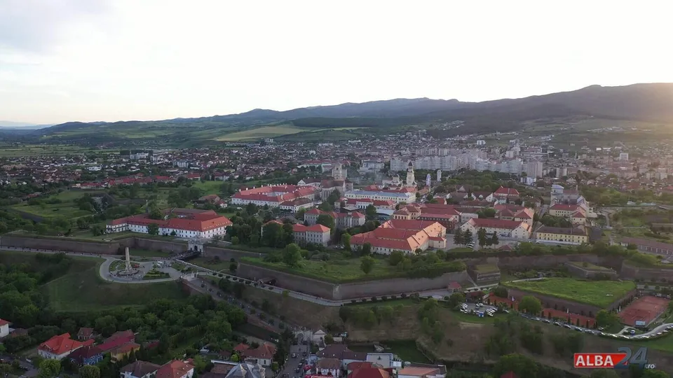 O vedere aeriană panoramică asupra unui oraș cu clădiri cu acoperișuri roșii, dealuri verzi și munți în fundal sub un cer deschis.