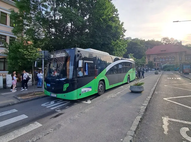 Un autobuz verde și negru cu numărul 20 este oprit pe o stradă lângă un trotuar, în apropierea unor persoane și a unor copaci.