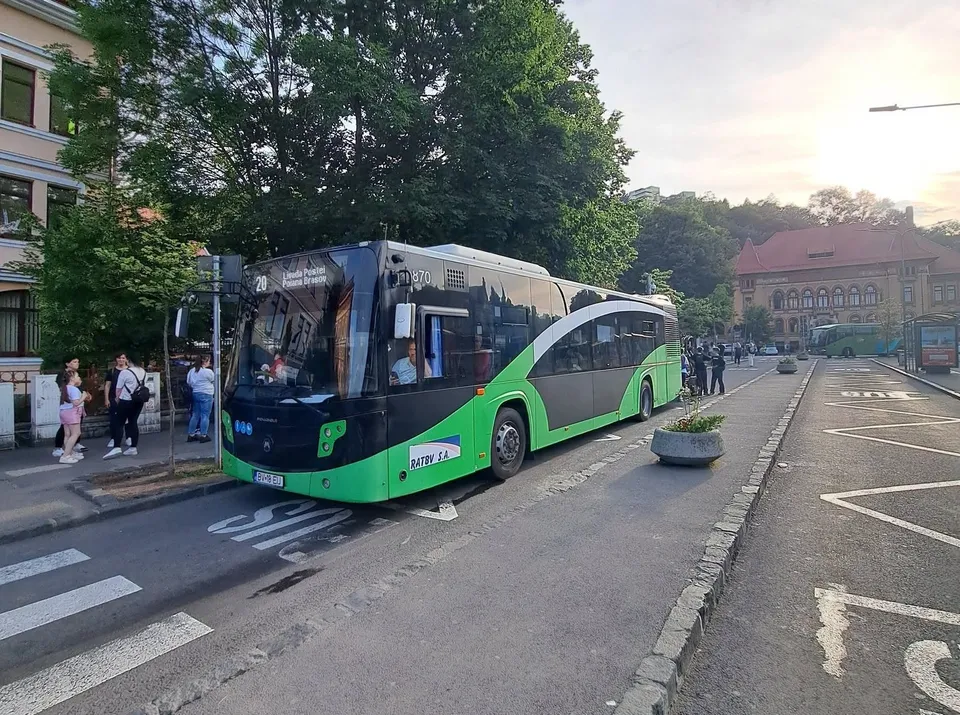 Un autobuz verde și negru cu numărul 20 este oprit pe o stradă lângă un trotuar, în apropierea unor persoane și a unor copaci.