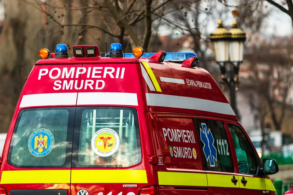Partea din spate a unei ambulanțe roșii cu inscripții albe și galbene, dotată cu girofaruri colorate pe plafon.