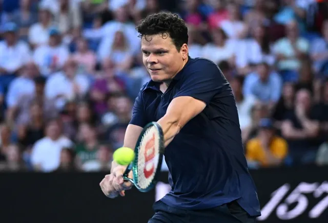 Un bărbat cu tricou închis la culoare ține o rachetă de tenis și privește spre o minge galbenă în timpul unui meci.