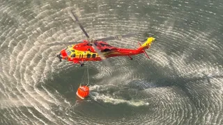 Un elicopter roșu și galben zboară deasupra unei suprafețe de apă, transportând un recipient portocaliu suspendat prin cabluri.
