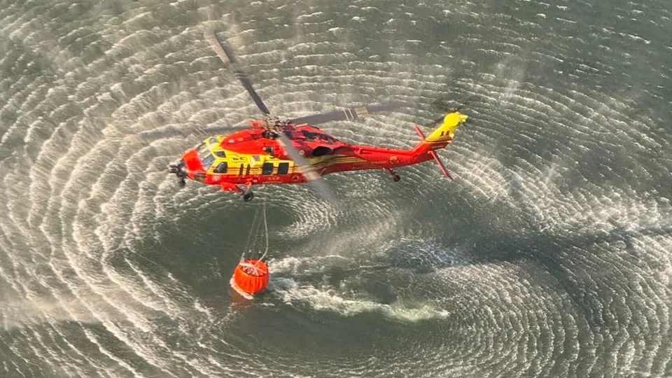 Un elicopter roșu și galben zboară deasupra unei suprafețe de apă, transportând un recipient portocaliu suspendat prin cabluri.