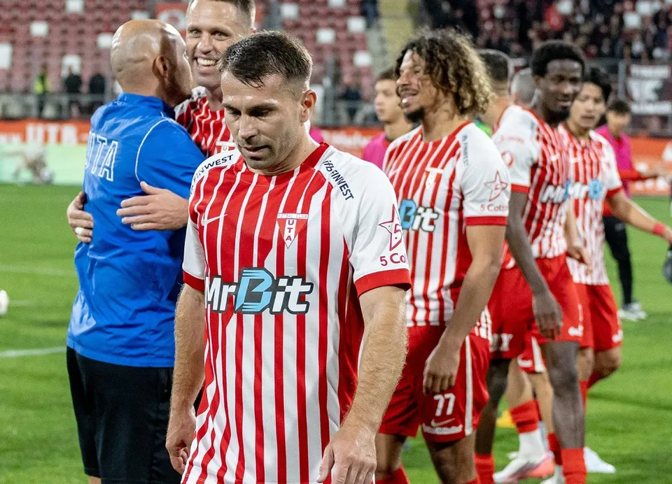 Un grup de fotbaliști în echipamente de joc cu dungi roșii și albe stau pe un teren de fotbal, în timp ce un bărbat în blu de antrenor îi îmbrățișează.