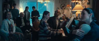 Un grup de persoane stă într-o încăpere luminată slab, cu un individ în prim-plan ținând un obiect rotund și o lampă aprinsă pe fundal.