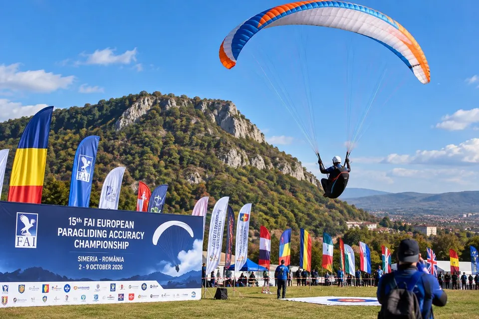 Un parapantist zboară sub o canopă albastră, portocalie și albă deasupra unui câmp cu steaguri și un panou informativ colorat.