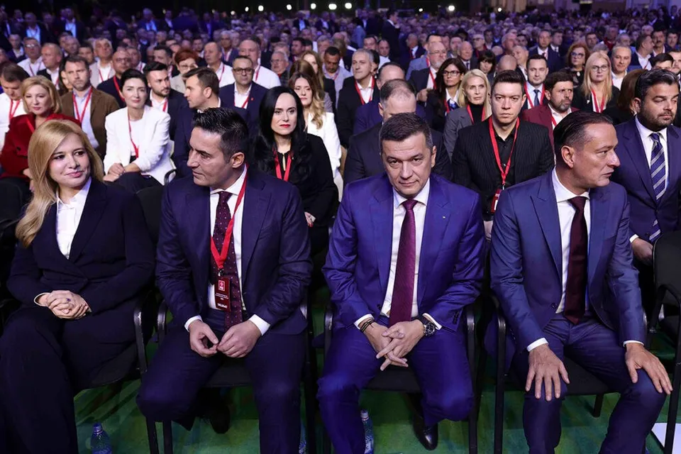 Un grup de persoane în haine formale stă așezat în rânduri de scaune într-o sală de conferințe plină de oameni.
