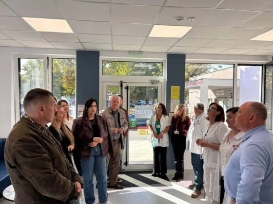 Un grup de persoane în haine civile și uniforme albe de medic stau în picioare într-o încăpere cu uși de sticlă și ferestre mari.