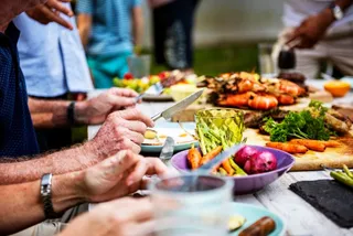 Oameni stau la o masă exterioară și consumă diverse alimente, inclusiv legume, pe farfurii și într-un bol colorat.