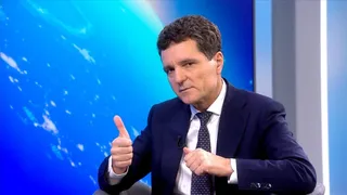 Un bărbat cu părul scurt și costum închis, cămașă albă și cravată cu puncte, face gestul cu degetul mare ridicat în fața unui fundal albastru.