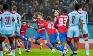 Câțiva jucători de fotbal în echipamente roșii și albastre sărbătoresc pe un teren de fotbal în fața unui grup de spectatori.