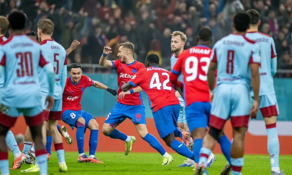 Câțiva jucători de fotbal în echipamente roșii și albastre sărbătoresc pe un teren de fotbal în fața unui grup de spectatori.