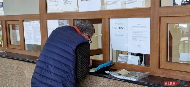 Un bărbat cu vestă albastră și ochelari privește documente afișate într-o vitrină de lemn cu geamuri.
