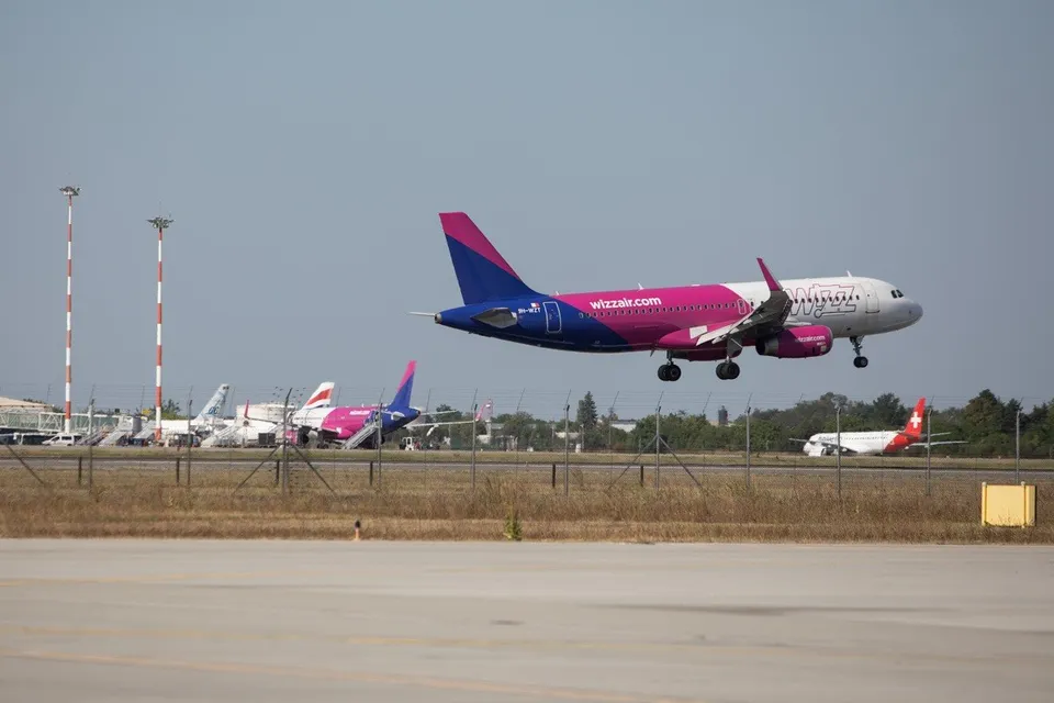 Un avion de pasageri de culoare roz și albă zboară deasupra unei piste de aterizare, având alte aeronave parcate în fundal.