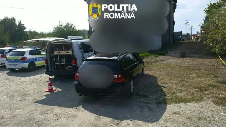 Două mașini de poliție și un autoturism negru sunt parcate pe un teren de pietriș lângă o clădire și vegetație.
