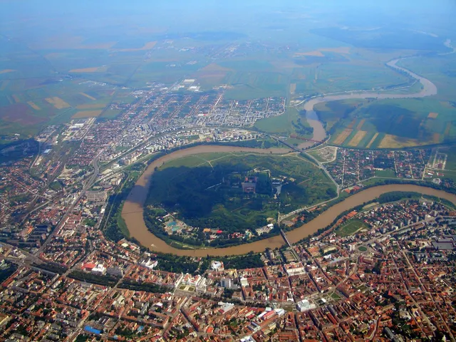 O vedere aeriană a unui oraș cu clădiri, străzi și un râu care curge prin zona urbană și câmpie.