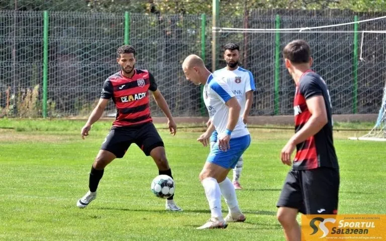 Trei bărbați joacă fotbal pe un teren de iarbă verde, unul având un balon la picioare, în fața unui gard de sârmă.