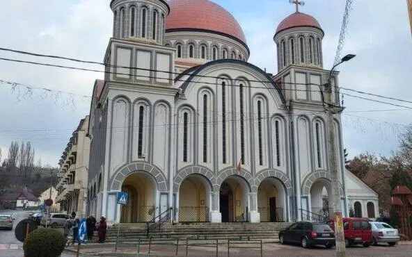O clădire de biserică ortodoxă cu un cupolă mare roșie și turnuri mai mici, cu arce străpunse pe fațadă și un grup de oameni în picioare lângă intrare.
