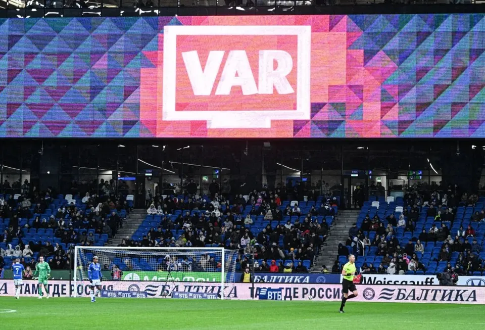 Un ecran mare afișează textul VAR pe un fundal colorat, deasupra unui stadion cu spectatori așezați pe tribunele albastre și jucători pe teren.