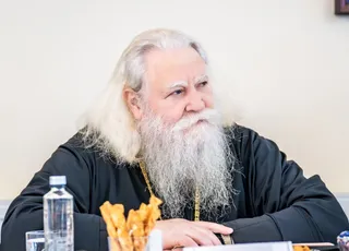 Un bărbat cu părul lung și alb și barbă lungă poartă o vestă neagră cu lanț auriu, stând la o masă cu o sticlă de apă.