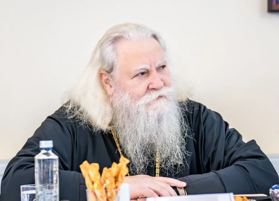 Un bărbat cu părul lung și alb și barbă lungă poartă o vestă neagră cu lanț auriu, stând la o masă cu o sticlă de apă.