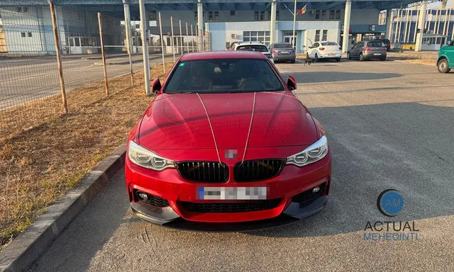 Un autoturism roșu este parcat pe un drum asfaltat în fața unei clădiri cu gard de sârmă și câteva vehicule în fundal.
