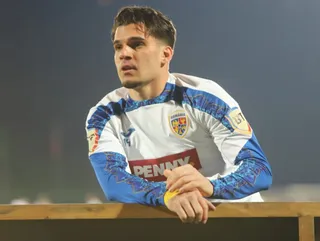 Un bărbat cu părul scurt poartă un tricou alb cu mâneci lungi albastre și se sprijină de o bordură de lemn.