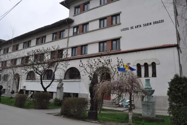 O clădire albă cu ferestre multiple, având inscripția Liceul de Artă Sabin Dragoi pe fațadă, este înconjurată de copaci și arbuști.
