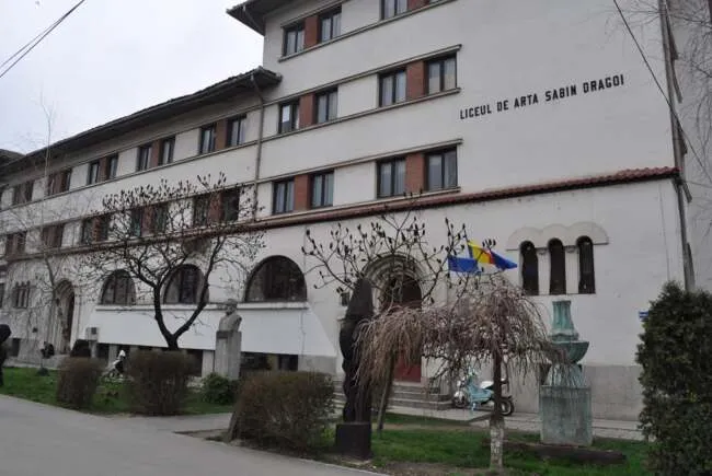 O clădire albă cu ferestre multiple, având inscripția Liceul de Artă Sabin Dragoi pe fațadă, este înconjurată de copaci și arbuști.