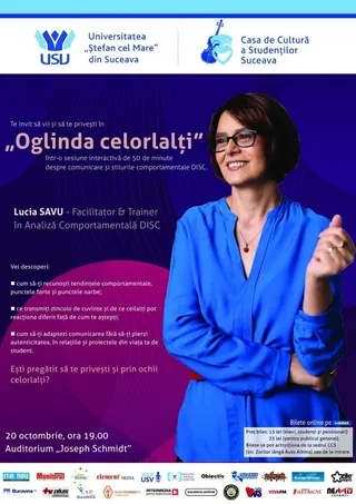 O femeie cu ochelari și bluză albastră stă în fața unui fundal violet pe care sunt scrise diverse texte și simboluri grafice.
