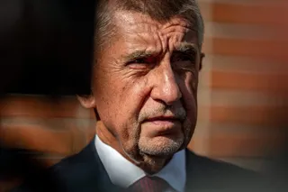 Un bărbat în vârstă, cu părul scurt și gri, poartă o cămașă albă, o cravată roșie și un sacou închis la culoare.